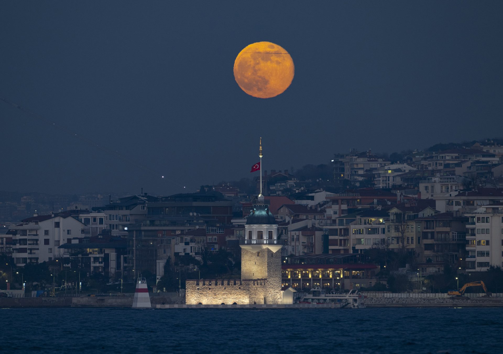 A glowing full moon rises above the historic İstanbul Kız Kulesi (Maiden's Tower) at night, with a backdrop of a densely built hillside and calm waters in the foreground.